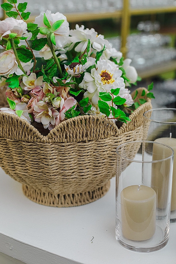 Wedding centerpiece with a floral centerpiece basket of white and blush flowers and greenery, plus glass hurricane vases with pillar candles on a white tabletop