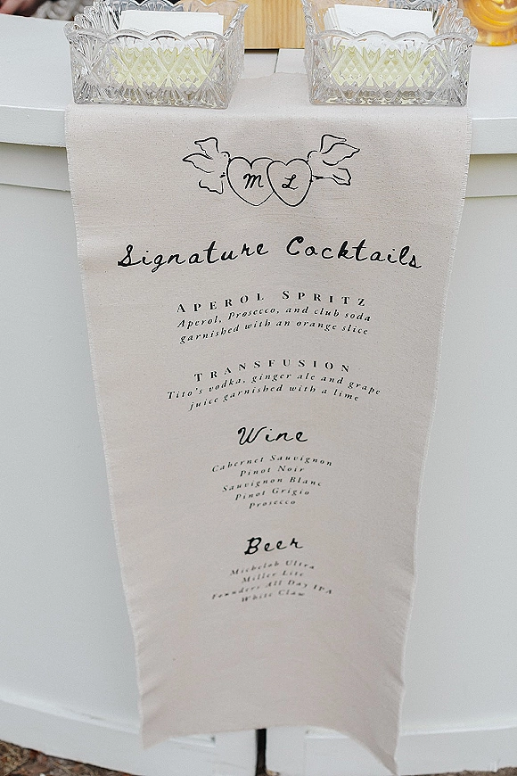 Signature cocktail sign displayed on a bar cart with crystal glass bowls and cocktail napkins on a white outdoor counter