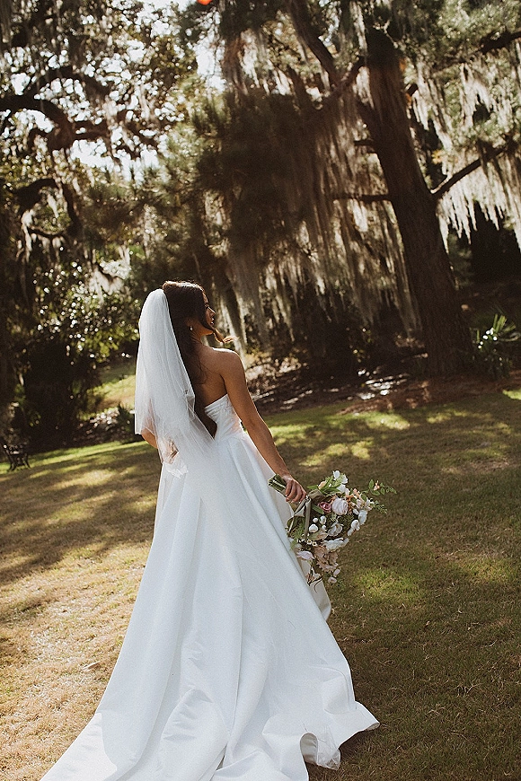 Bridal portrait of a bride walking away in a strapless ball gown with long veil and bouquet beneath oak trees with hanging moss