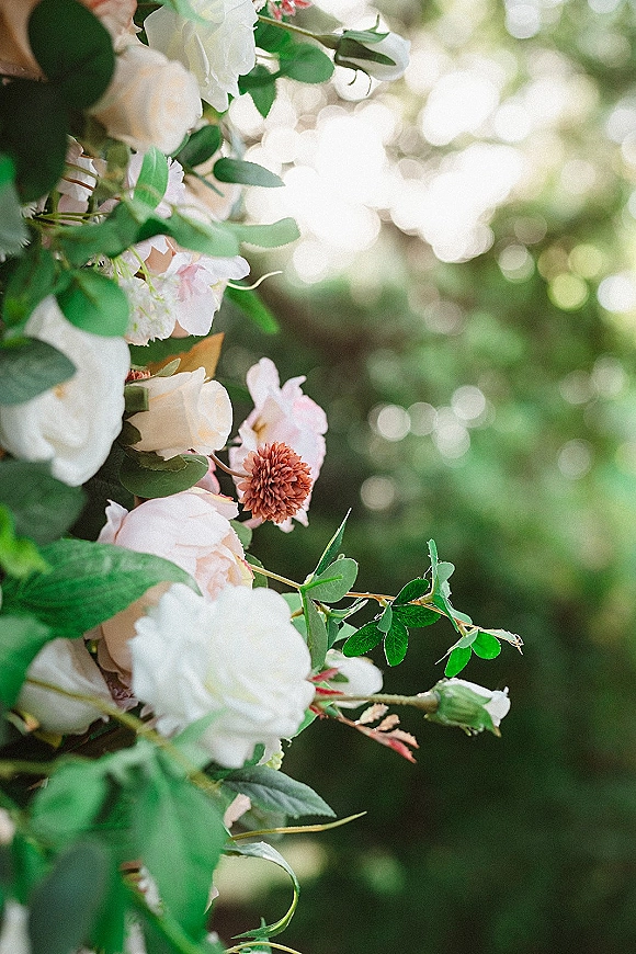Wedding floral arrangement with white roses, blush blooms, and greenery on a ceremony floral arch, set against sunlit trees with soft bokeh