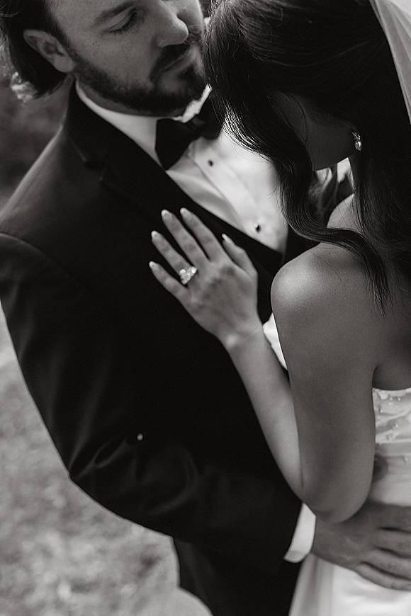 Wedding couple portrait in a black and white wedding photo, groom kissing bride’s forehead as she rests a ringed hand on his chest outdoors