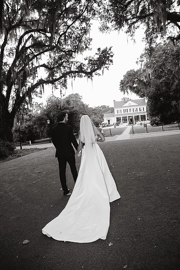 Wedding couple portrait of newlyweds holding hands, walking away down a path under tree canopy with Spanish moss, bride’s veil and long train trailing
