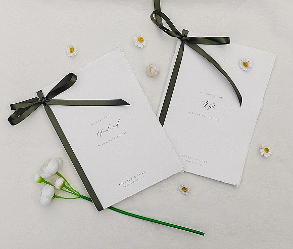 Wedding vow books with dark green ribbon bows and calligraphy on deckled edge paper, styled with small daisies on a white flatlay