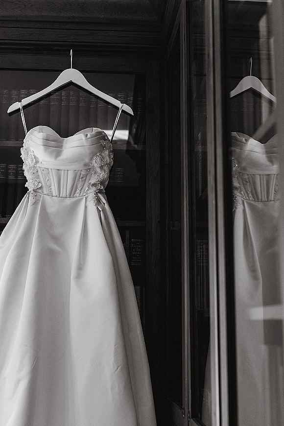 Wedding dress, strapless satin gown with corset bodice, floral appliques and beading on a hanger by bookshelves with reflection in glass doors