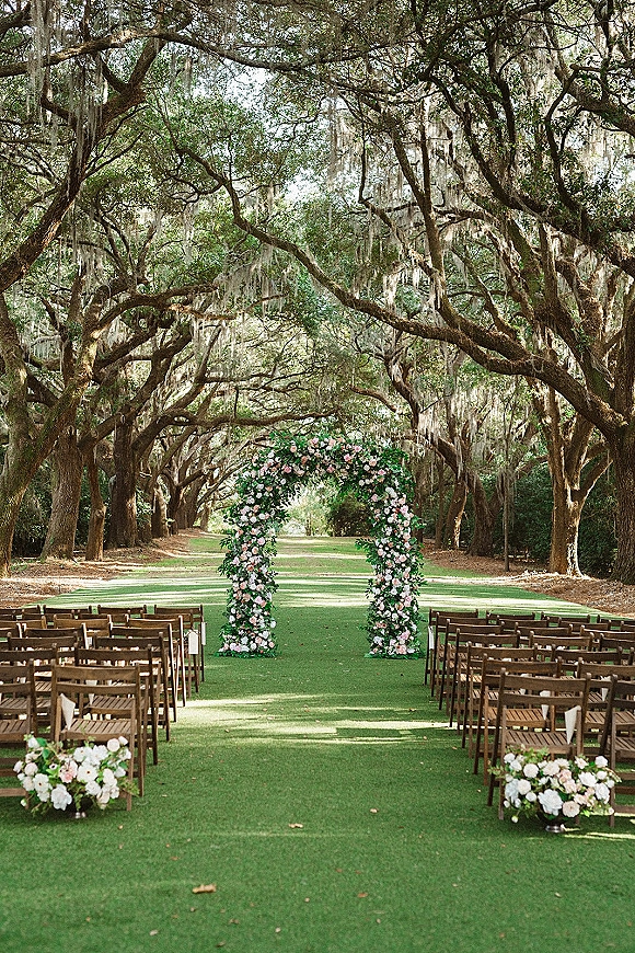 Ceremony setup for an outdoor wedding ceremony with a pink and white floral arch, wooden folding chairs, and aisle runner beneath mossy oak trees