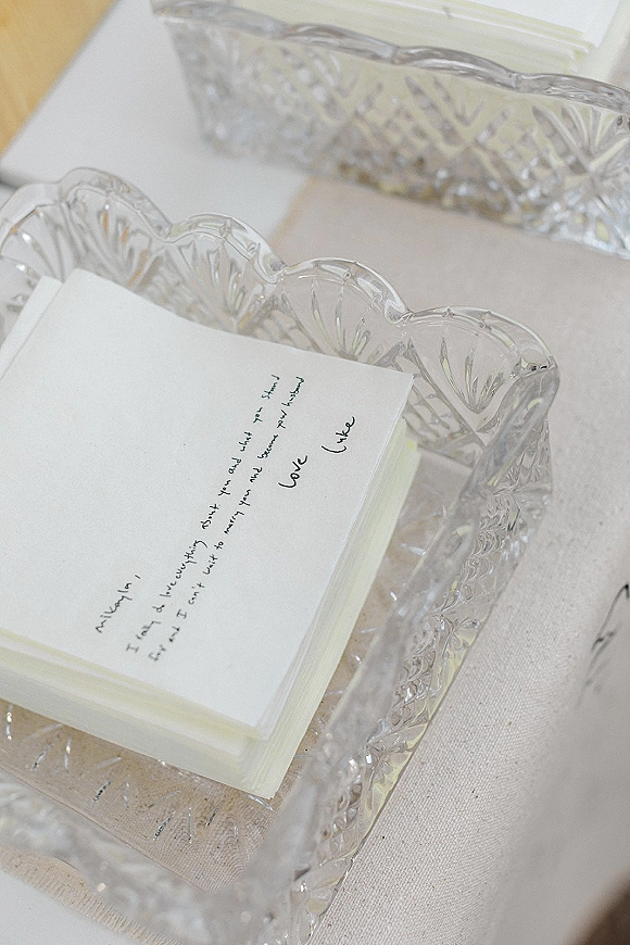 Wedding vow cards with handwritten wedding vows resting in a clear cut glass dish on a linen napkin atop a neutral tablecloth flat lay