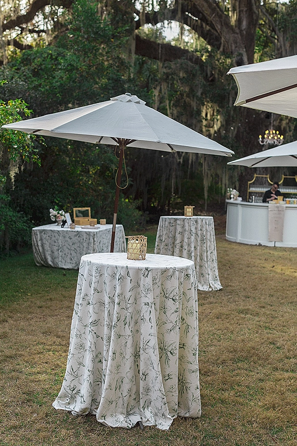 Cocktail hour setup with outdoor cocktail hour tables in patterned linens, umbrellas and lanterns beside a bar under trees with hanging moss
