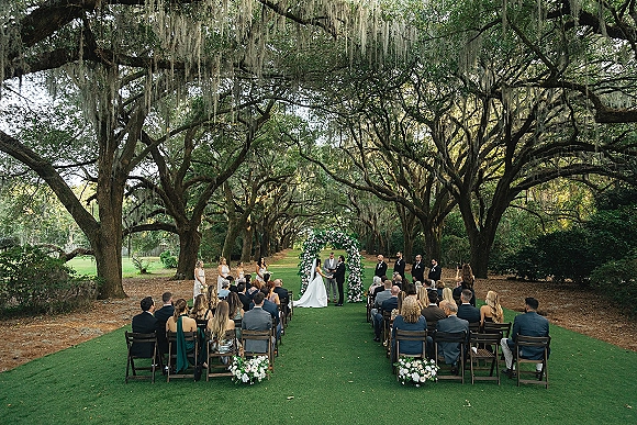 Outdoor wedding ceremony with a floral arch, bride and groom at the altar, and guests seated under oak trees draped in Spanish moss