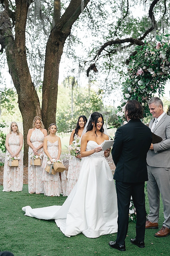 Wedding vows as bride reading vows from a book beside groom under a floral arch, cathedral veil flowing on lawn beneath oak trees and moss
