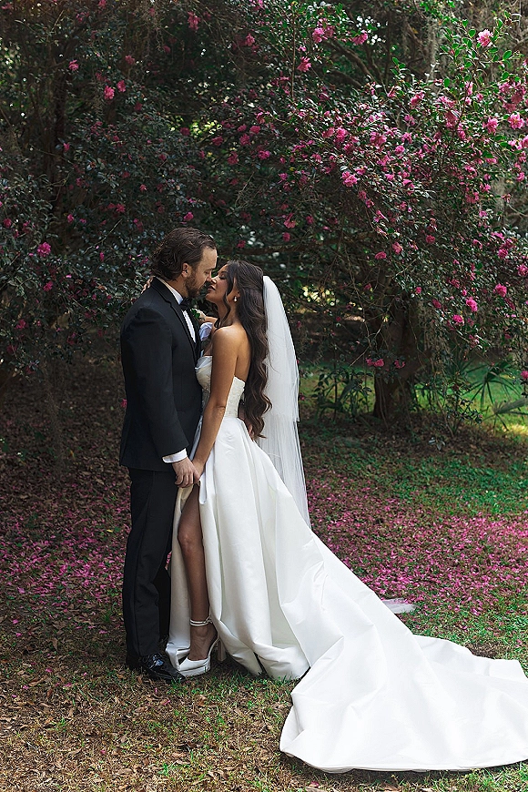 Wedding couple portrait of bride and groom kissing under a flowering tree with pink blossoms, her cathedral veil trailing over the lawn