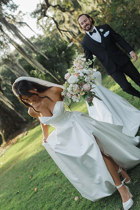 Couple portrait of bride in a strapless gown with bouquet and veil, groom in black tuxedo, walking on a mossy garden lawn