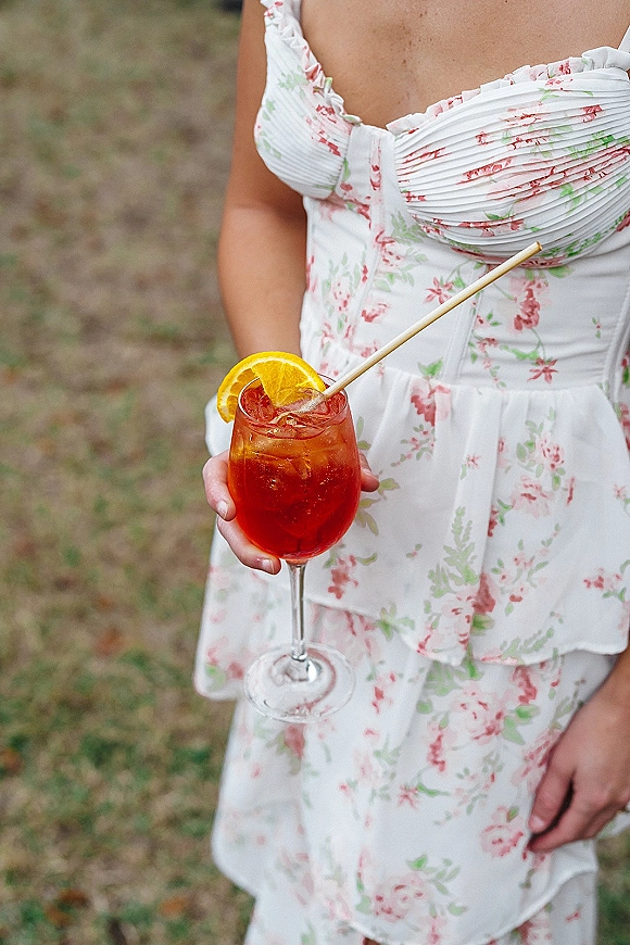 Wedding cocktail in a stemmed wine glass with red spritz, ice, and orange slice garnish, held by someone in a floral dress on grass lawn