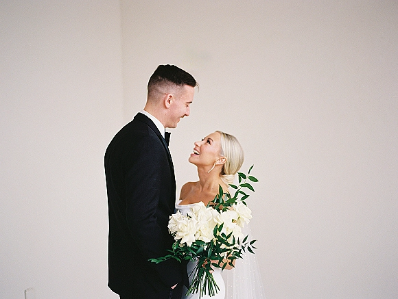 Couple portrait of bride looking up at groom, holding a white rose bouquet with greenery, posed against a minimalist white wall