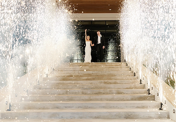 Wedding exit moment as bride and groom hold hands walking through cold spark fountains on a stone staircase at a modern entrance