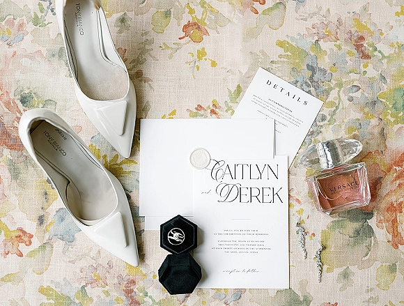 Invitation suite and wedding invitation flat lay with wax seal, envelope, white pointed-toe heels, ring and perfume on floral linen background