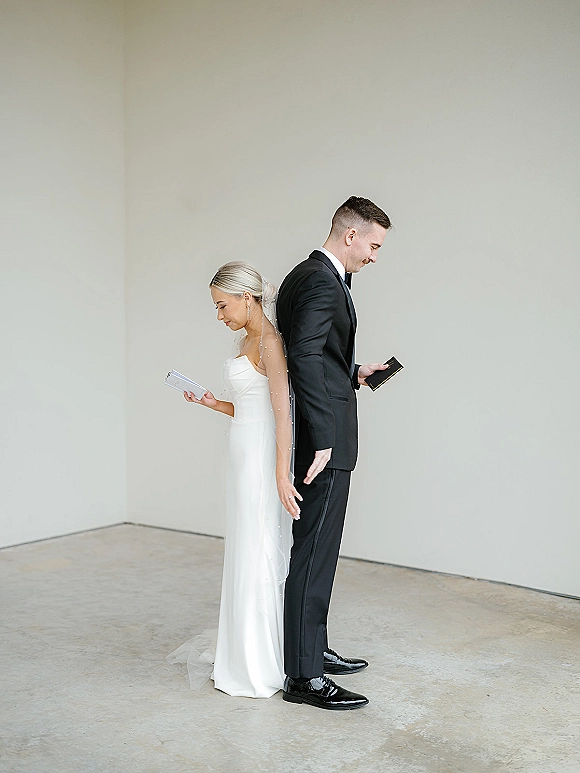 First look moment as bride and groom stand back to back holding vow books, no peeking, against a minimalist indoor wall on concrete floor