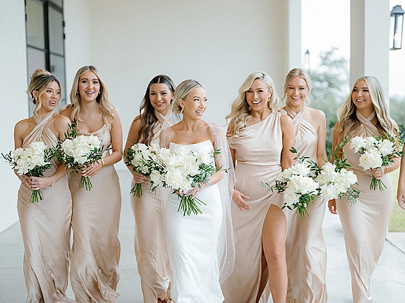 Bride with bridesmaids in a bridal party portrait, walking under a covered walkway with columns, holding white bouquets with greenery and veil flowing