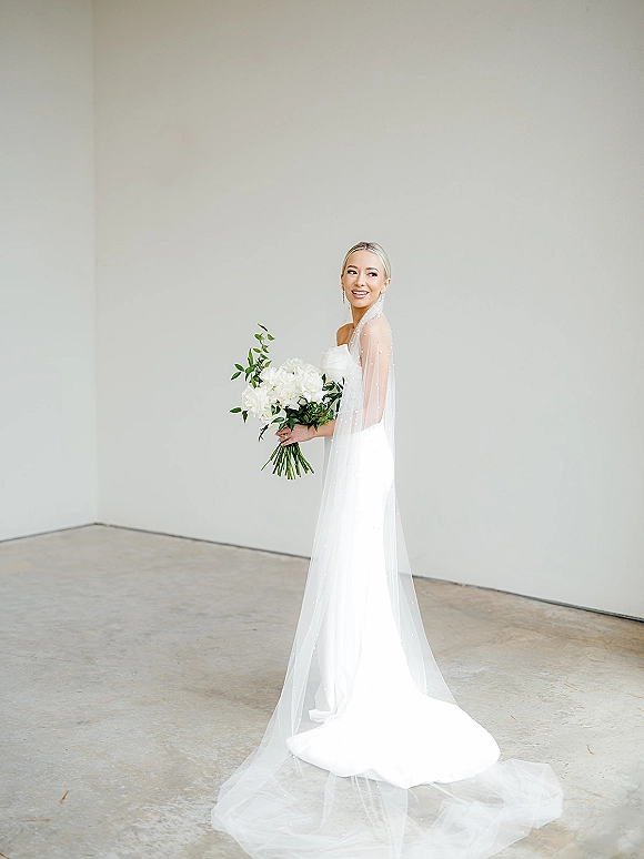 Bridal portrait of a bride in a simple wedding dress with long veil, holding a white flower bouquet in a minimalist studio setting