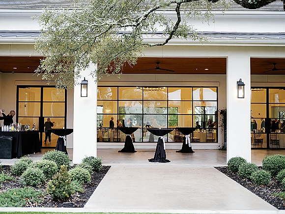Cocktail hour setup with wedding cocktail hour black linen high-top tables, bar with glassware, and string lights under a covered patio with grid windows