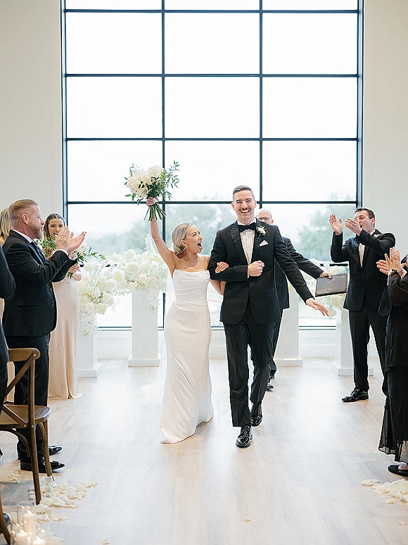 Wedding recessional as bride and groom walk the aisle, her bouquet raised, guests applauding in a bright modern indoor space with large grid window