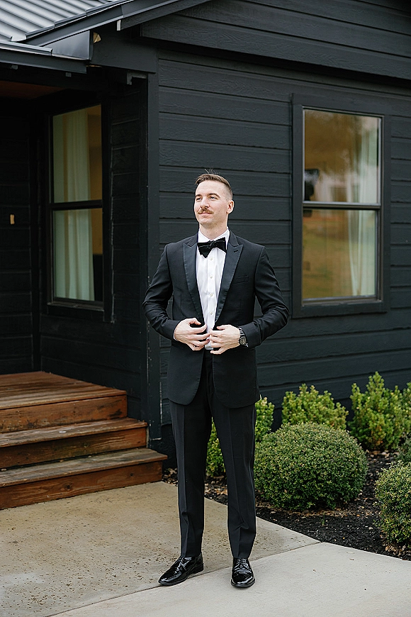 Groom portrait of a groom in tuxedo adjusting his black tuxedo jacket and bow tie on wooden steps outside a black house exterior