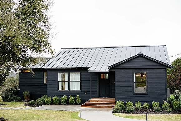 Wedding venue exterior of a modern barn venue with black siding, metal roof, wood steps, and lit entry framed by shrubs and lawn