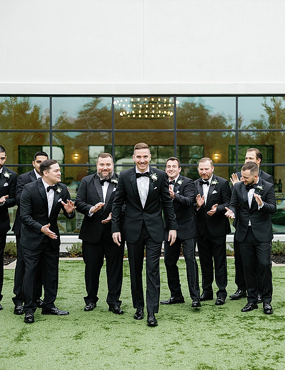 Groom with groomsmen group photo in matching black tuxedos and bow ties on a lawn outside a modern glass-front venue with reflections