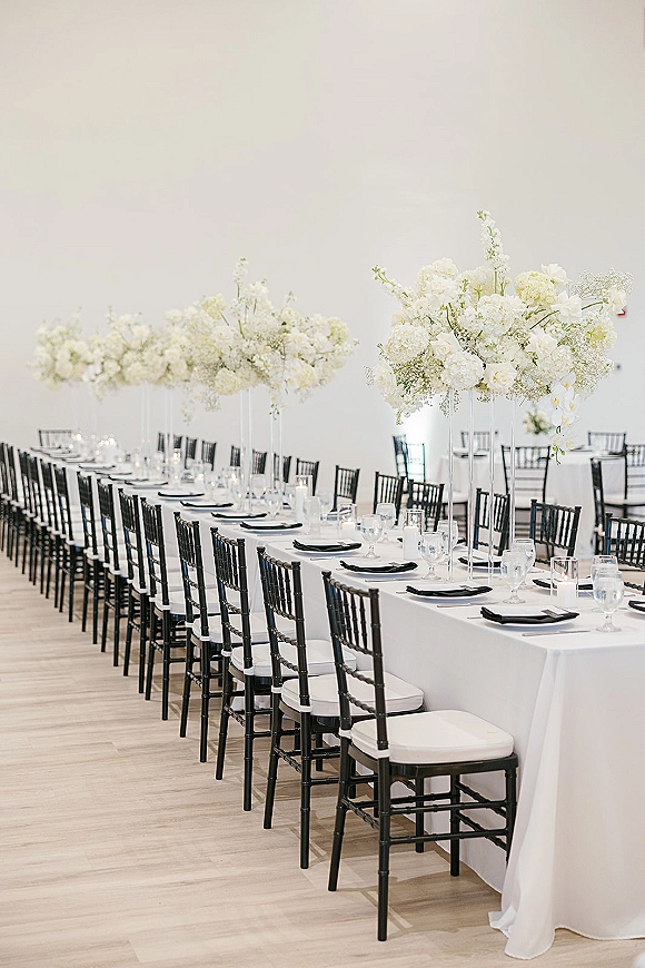 Reception tablescape with a long wedding banquet table dressed in white linen, black Chiavari chairs, tall white florals, and votive candles indoors