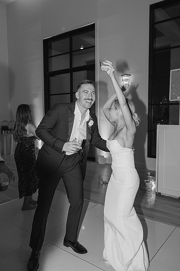 Wedding reception dancing as bride and groom sway with cocktail glasses, her strapless gown and his tux, guests by tall windows behind