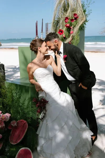 Couple portrait as the bride and groom touch foreheads on a sunlit beach, the groom's calla lily boutonniere bright against ocean and blue sky.