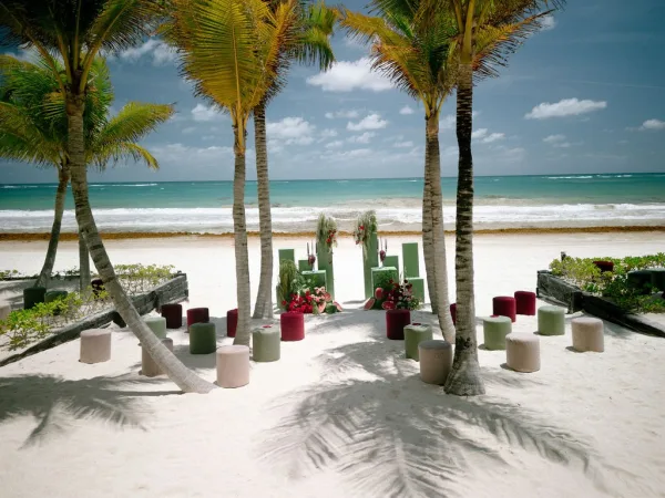 Beach ceremony setup with tropical ceremony decor and pouf seating, set on sand beside the ocean and framed by palm trees.