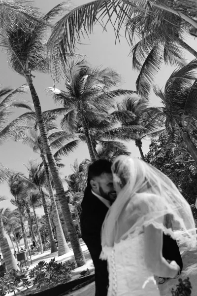 Couple portrait of a beach wedding couple embracing, her bridal veil catching the breeze beneath palm trees on a sandy beach.