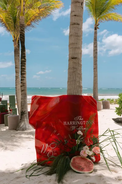 Wedding welcome sign, a piece of beach wedding signage, accented with a single anthurium, set on beach sand with ocean and palm trees beyond.