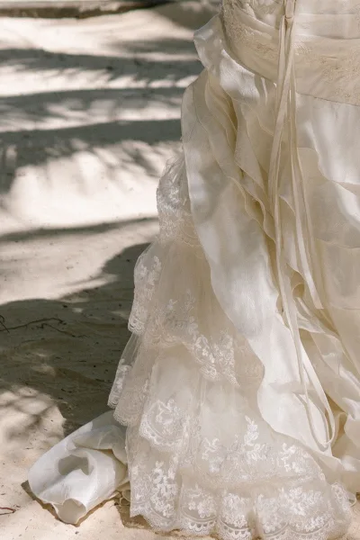 Wedding dress detail of a lace bridal gown with delicate lace appliqué, the tiered skirt brushing sand on a quiet beach at golden hour.