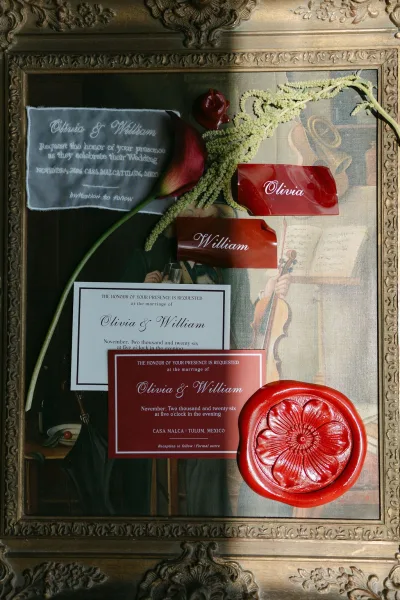 Wedding invitation suite featuring burgundy wedding invitations and a wax seal, framed in an ornate gold frame before a vintage painting.
