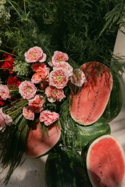 Wedding florals styled with a playful watermelon accent, arranged amid green foliage with soft shadows across a clean white floor.
