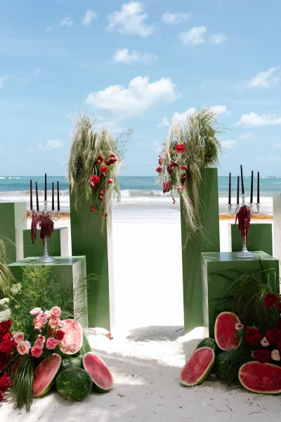 Beach ceremony decor for a modern wedding altar styled with watermelons, set on white sand before a blue ocean and blue sky.