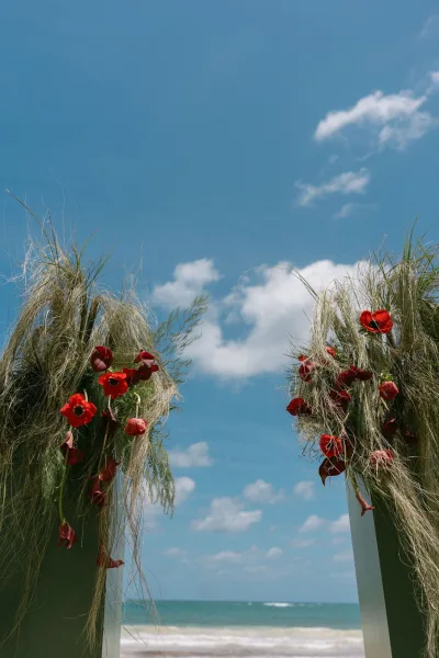 Beach wedding altar with red flowers forms an oceanfront ceremony backdrop on a sandy shore beneath blue sky and soft clouds.
