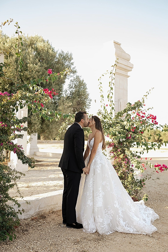 Wedding kiss as bride and groom hold hands under a floral arch with pink bougainvillea and white columns in a garden setting