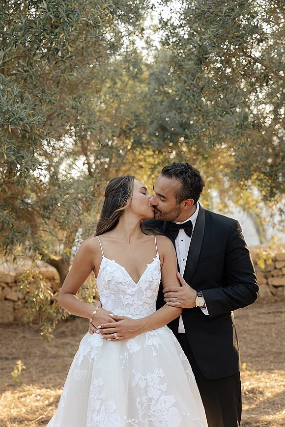 Wedding kiss portrait of bride and groom kissing, groom hugging from behind, bride in lace dress, sunlight with trees and stone wall backdrop