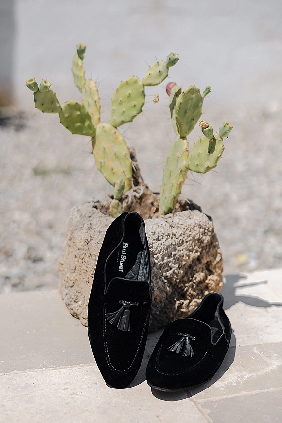 Groom shoes, black wedding loafers with tassels beside a cactus on a stone patio, with sand and open sky in the background