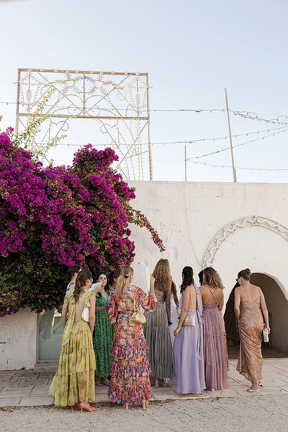 Wedding guest group in colorful maxi dresses chatting by a white stucco arched doorway in a courtyard with bougainvillea flowers overhead