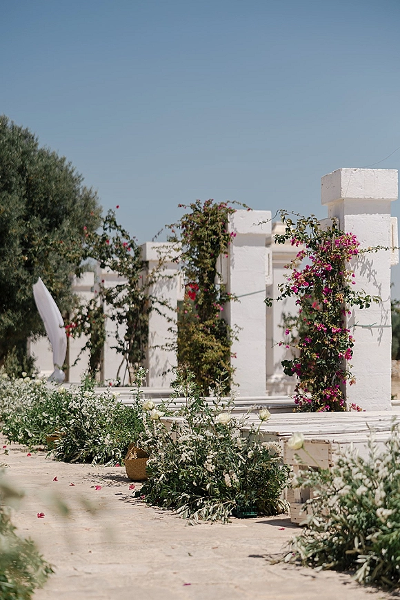 Ceremony aisle decor with an outdoor wedding aisle lined in greenery garlands, white flowers, and rose petals on a stone path under blue sky