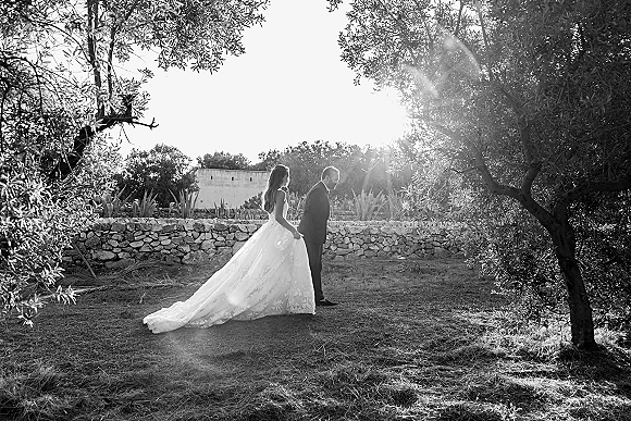 Couple portrait in a black and white wedding portrait, bride and groom walking with her long train on grass by a stone wall and trees