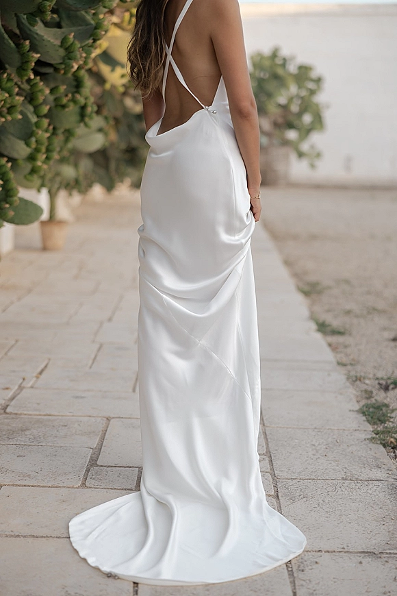 Wedding dress with a low back satin gown and spaghetti straps, small train trailing on a stone walkway beside potted greenery and a white wall