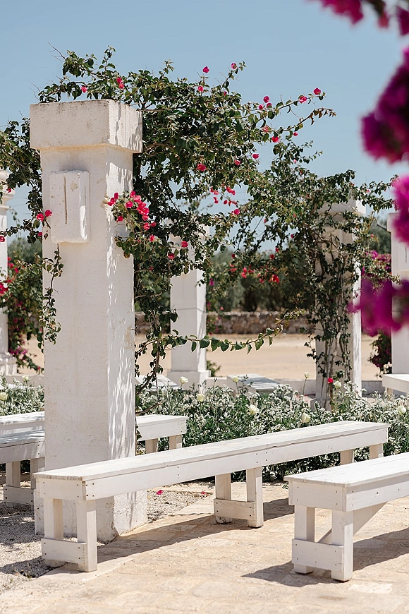 Outdoor ceremony setup with white wooden benches and vine-wrapped columns, pink and white flowers, under a blue sky beside a stone wall