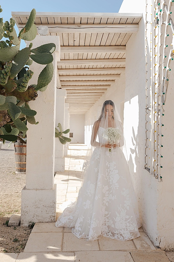 Bridal portrait of a bride holding bouquet, looking down in a strapless ball gown with long veil in a white stucco corridor with string lights