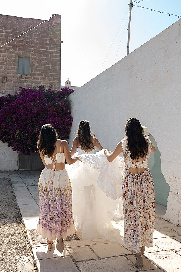 Bride with bridesmaids walking to ceremony as they lift her long train, backs turned on a paved walkway by a white stucco wall with bougainvillea accents