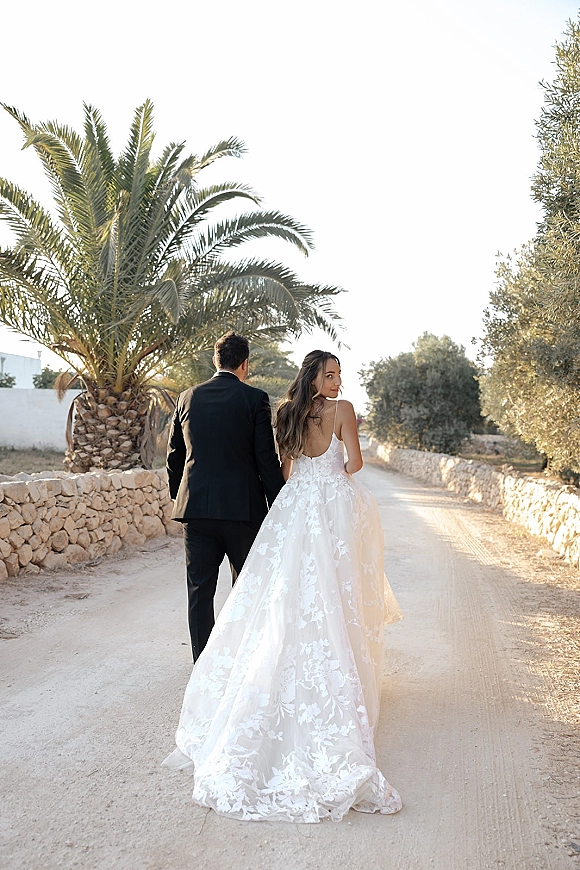 Couple portrait of bride and groom walking away holding hands, bride looking back, lace train and veil on a dirt road by a stone wall