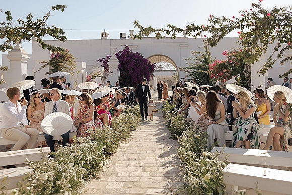 Wedding processional with flower girl walking down aisle holding a basket, guests under white parasols in a sunny courtyard gate setting